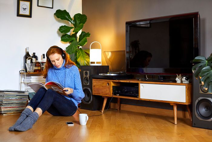A woman in a blue turtleneck sweater sits on the wood floor in front of her TV and stereo. She is wearing over ear headphones that are plugged in to the stereo as she reads a magazine.